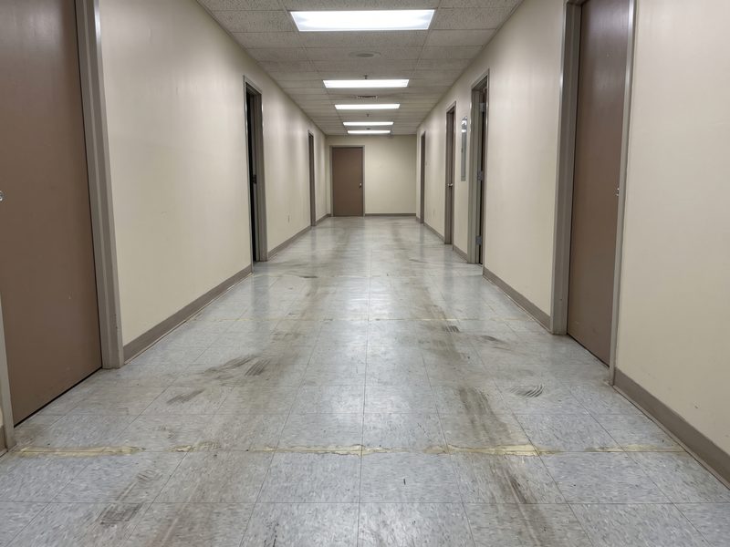 A commercial corridor vinyl tile floor with dull worn wax, black heel scuffs and dark walked-in traffic lanes, before stripping and waxing