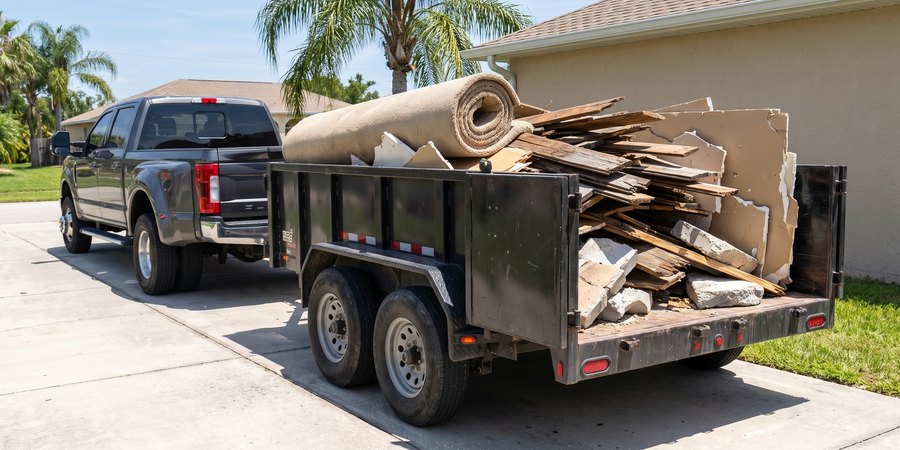 A dump trailer loaded with torn-out decking, drywall and rolled carpet, hitched to a pickup on a Florida driveway